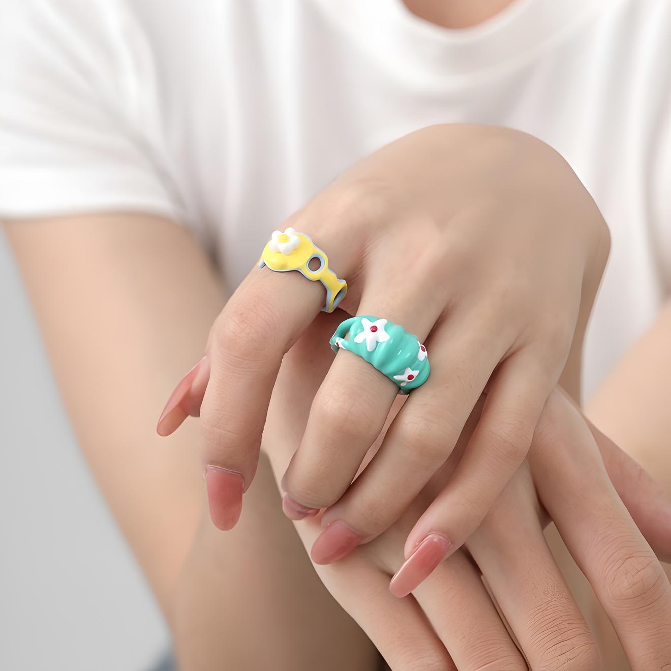 Colorful enamel rings with playful designs worn on a woman's fingers, showcasing fun fashion accessories with a white t-shirt backdrop.