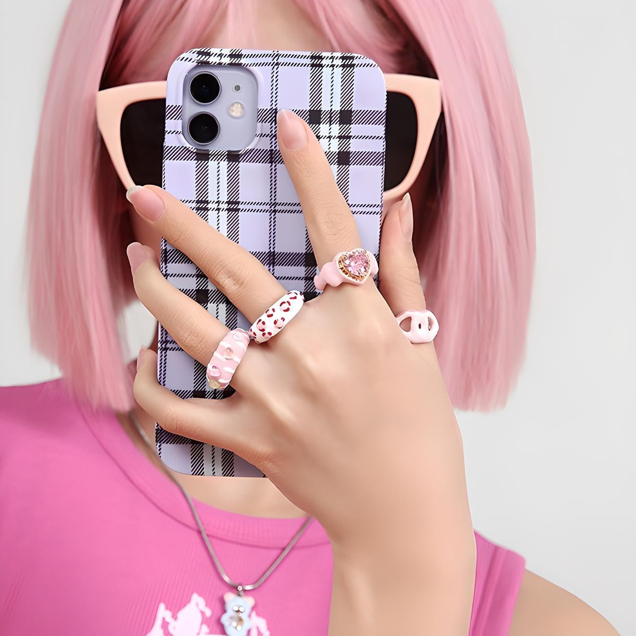 Fashionable woman with pink hair flaunting stylish plaid phone case and trendy rings, coordinating with her vibrant pink outfit for a chic look.