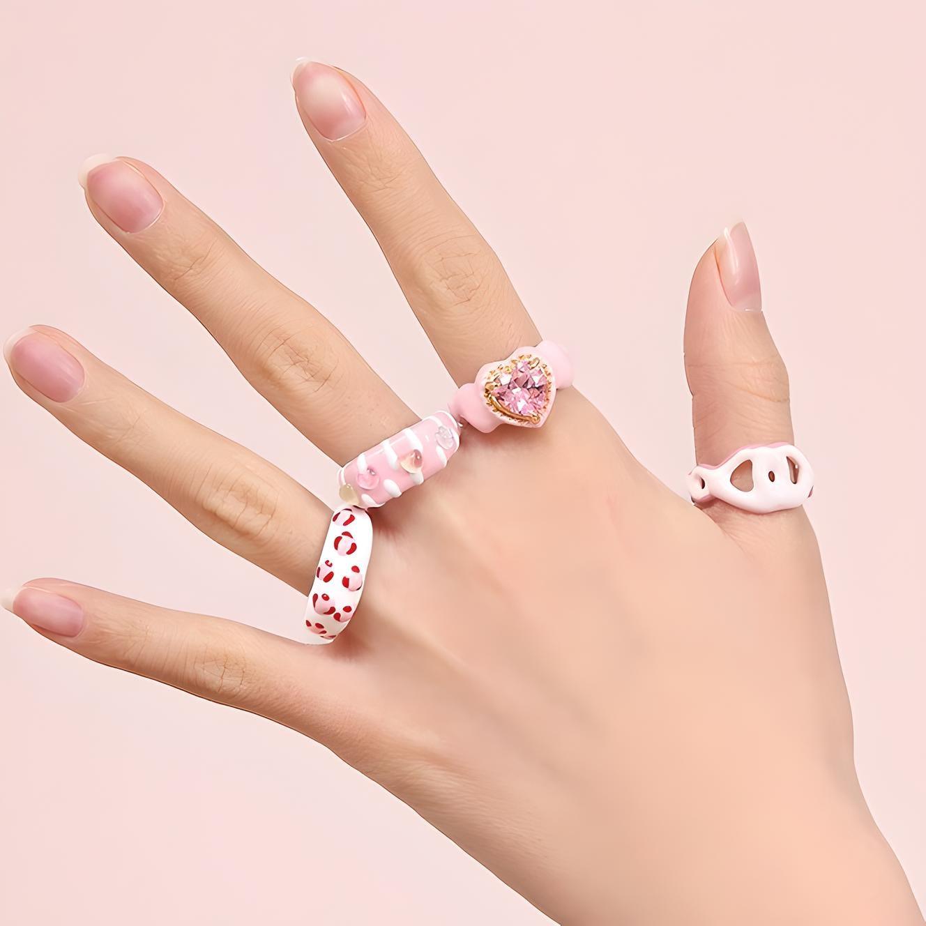 Elegant hand displaying trendy rings with pastel tones and playful designs, on a soft pink background – perfect for a chic, youthful style statement.