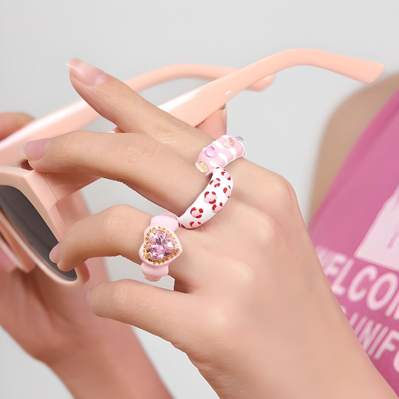 Close-up of elegant hand with stylish nail art, adorned with heart-themed rings, holding a pink smartphone case. Chic fashion accessories perfect for a trendy outfit.