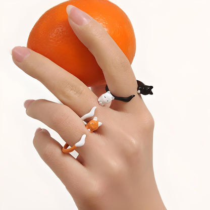 Elegant hand with glossy manicure showcases quirky cat rings while gripping a vibrant orange, offering a playful yet sophisticated jewelry style. Perfect for cat lovers looking to add whimsy to their accessories.