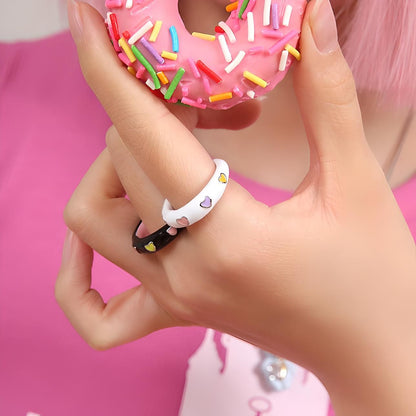 Close-up of a pink-frosted donut with sprinkles being held by a person wearing stylish heart-patterned rings, against a pink background—ideal for fashion and foodie-themed content.